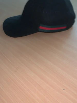 Svart Gucci keps med röd-grön rand - Snygg svart keps från Gucci med klassiskt GG-mönster i tyget och en ikonisk röd-grön rand på sidan. Kepsen har böjd skärm och justerbar passform. Materialet är textil som känns både lätt och bekvämt på huvudet.