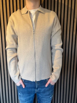 Beige stickad tröja med dragkedja från Sandö - Beige stickad tröja med krage och hel dragkedja framtill. Tröjan har lång ärm och en clean, minimalistisk look som passar till jeans eller andra byxor. Perfekt för dig som gillar enkel och stilren design. Tröjan är från Sandö, skicka dm vid funderingar!