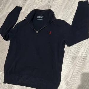Mörkblå stickad tröja Polo Ralph Lauren - Mörkblå stickad tröja från Polo Ralph Lauren med halv dragkedja och ribbad krage. Klassisk röd broderad logga på bröstet. Långärmad och tillverkad i bomull, perfekt för lager-på-lager under kyliga dagar. Inga hål eller liknande 