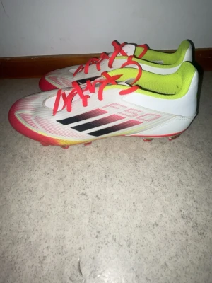 Adidas F50 vita fotbollsskor - Säljer ett par Adidas F50 fotbollsskor i vitt med svarta och röda detaljer. Skorna har röda snören, neongul insida och klassiska tre svarta ränder på sidan. Ovandelen är i syntetmaterial och sulan har dobbar för spel på gräs. Perfekta för dig som vill ha lätta och snabba skor.