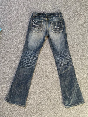Blå bootcut jeans med nitar från Gudanlu - Säljer ett par jätte coola och unika blå bootcut jeans från Gudanlu med slitningar och coola nitar på fickorna. Baksidan har en stjärndetalj och text på ena fickan. Jeansen har låg midja och är tillverkade i denim med stretch. Säljer för att de inte kommer till användning💞