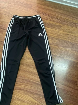 Svarta Adidas mjukisbyxor med vita ränder - Säljer ett par svarta mjukisbyxor från Adidas med klassiska vita ränder längs benen och logga på låret. Byxorna har dragsko i midjan och dragkedja vid bensluten. Perfekta för chill eller träning.