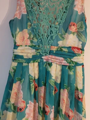 Blommig långklänning med spetsdetalj - Långklänning i turkosblått med stora blommor i rosa, vitt och grönt. Djup v-ringning med dekorativ spets i samma färg som klänningen. Markerad midja och svepande kjol. Perfekt för sommarens alla tillfällen.