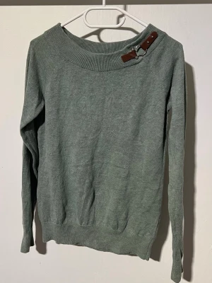Grön stickad tröja med läderdetalj - Snygg grön stickad tröja med lång ärm och bred ribbad krage. Unik detalj med brun läderrem och metallspänne vid halsen som ger en cool touch. Perfekt för dig som gillar stilrena plagg med något extra. Materialet känns mjukt och bekvämt.