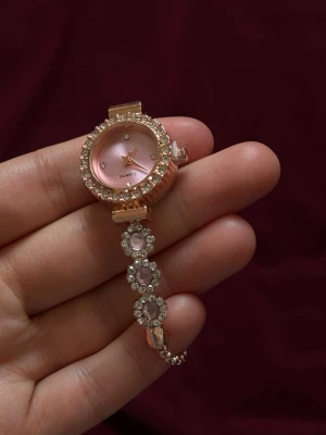 Rosa glittrig klocka med stenar - Säljer en elegant rosa klocka med glittrande stenar runt urtavlan och på armbandet. Klockan har en rund form, roséguldfärgad metall och gnistrande detaljer som ger en lyxig känsla. Perfekt accessoar för att lyfta din stil.