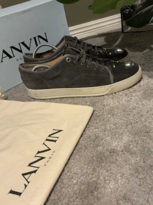 Gråa Lanvin sneakers i mocka och lack - Snygga grå sneakers från Lanvin med ovandel i mocka och detaljer i svart lack. Skorna har vita sulor och klassisk låg profil med snörning. Perfekta för dig som gillar stilrena och exklusiva sneakers med en lyxig touch.