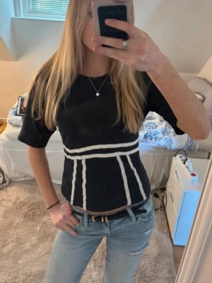 Svart t-shirt med vita detaljer - Snygg svart t-shirt från Gina med vita ränder och coola detaljer i midjan. T-shirten har korta ärmar och öppen rygg med knyt baktill. Passformen är normal och materialet känns som bomull. 💓💓Endast använd 2 gånger (lånade bilder) Sista bilden är egen och visar en svart fläck av annan nyans av tröjan (upplever inte att den syns på)