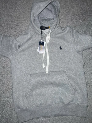 Grå hoodie från Polo Ralph Lauren - Snygg grå hoodie från Polo Ralph Lauren med vit dragsko i huvan och klassisk känguruficka. Broderad marinblå logga på bröstet. Tillverkad i mjuk bomullsblandning för skön känsla. Perfekt för en avslappnad och stilren look.