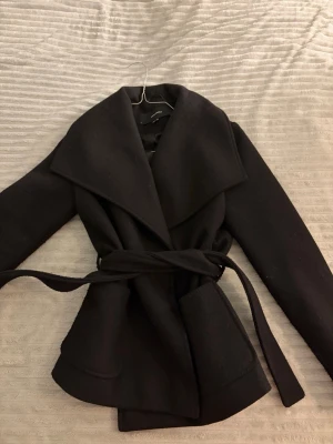 Svart kappa från Vero Moda med skärp - Stilren svart kappa från Vero Moda med bred krage och knytband i midjan. Kappan har två stora fickor framtill och är fodrad för extra komfort. Perfekt för kyliga dagar och ger en snygg siluett. Materialet är en mjuk ullblandning.