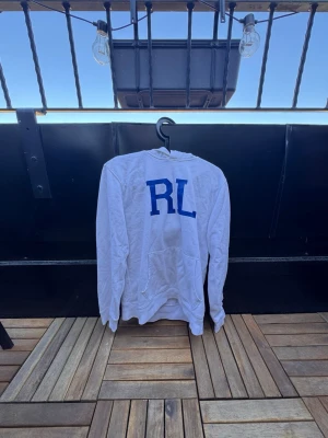 Vit hoodie från Ralph Lauren med  blå RL-tryck🙌🤩 - Säljer en vit hoodie med stora blå bokstäver 'RL' på bröstet från Ralph Lauren Hoodien har en klassisk huva och en stor magficka framtill. Tillverkad i mjukt material som känns skönt mot huden. Perfekt för dig som gillar enkel och stilren streetwear.🙌🤩
