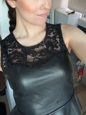 Svart tröja  med spetsdetaljer - Snygg svart klänning med ärmlös design och spets upptill. Klänningen har en rund halsringning och materialet ser ut som syntet med en glansig finish. Spetsen ger en elegant touch och gör plagget extra speciellt.