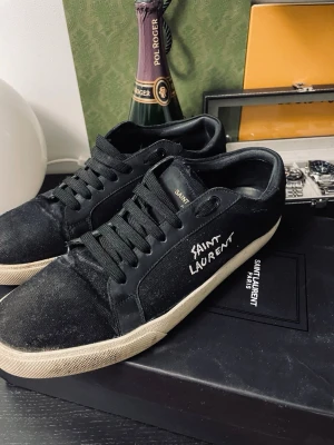 Svarta Saint Laurent sneakers - Svarta sneakers från Saint Laurent med vit logga på sidan och svart textil samt läderdetaljer. Klassisk låg modell med svarta skosnören och vit sula. Perfekta för dig som gillar stilrena och exklusiva sneakers med en edgy touch.