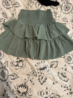 Grön volangkjol med smock - Supersöt grön kjol med bred smock i midjan och dubbla volanger. Kjolen är gjord i ett tunt, lätt tyg som ger ett flowy intryck. Perfekt för dig som gillar en feminin och trendig stil. Märke och material är okänt.