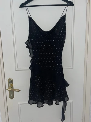 Svart festklänning med volang och strass - Snygg svart kortklänning med tunna axelband och små rhinestones. Klänningen har volangdetaljer längs sidan och nederkanten samt asymmetrisk fåll. Perfekt för dig som vill ha en unik och trendig look.🖤