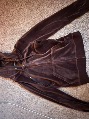 Brun velour hoodie från Juicy Couture - Mjuk och lyxig brun velour hoodie från Juicy Couture med dragkedja framtill, fickor och justerbar huva med snören. Klassisk logga på bröstet och ribbade muddar vid ärmslut och nederkant. Perfekt för en chill och trendig look.