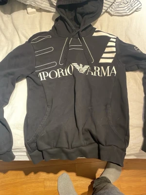 Svart hoodie från Emporio Armani - Snygg svart hoodie från Emporio Armani med stor logga och grafiskt tryck på bröstet samt EA7-detalj på ärmen. Hoodien har huva med dragsko och känguruficka framtill. Tillverkad i mjukt material, perfekt för en avslappnad streetwear-look.