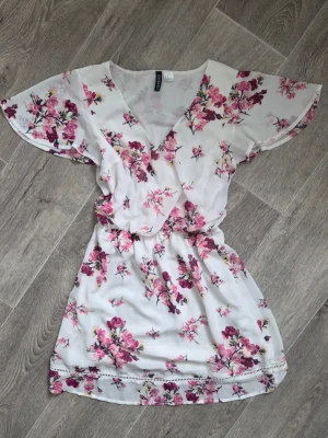 Vit blommig omlottklänning H&M - Söt kortklänning från H&M i vit med rosa blommönster. Fjärils-ärmar och resår i midjan. Tillverkad i ett lätt och luftigt polyestertyg. Har en fläck som jag ej provat få bort, se bild.