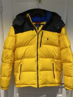 Gul pufferjacka från Polo Ralph Lauren - Gul pufferjacka från Polo Ralph Lauren med svarta detaljer upptill och på dragkedjorna. Jackan har hög krage, tre fickor med dragkedja och den klassiska Polo-loggan broderad på bröstet. Perfekt för kalla vinterdagar och riktigt snygg streetstil.