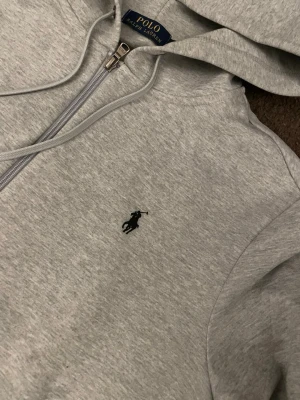 Grå hoodie från Polo Ralph Lauren - Snygg grå hoodie från Polo Ralph Lauren med dragkedja och huva. Klassisk design med logotyp på dragkedjan och mjukt material som känns riktigt skönt. Perfekt för dig som gillar stilrena och bekväma plagg.