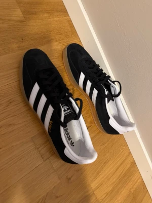  shoes Adidas Gazelle svart/vit - Säljer ett par klassiska Adidas Gazelle sneakers i svart mocka med vita detaljer och gummisula. Skorna har de ikoniska tre ränderna på sidan och texten 'GAZELLE' i guld. Låg modell med vit häl och svart fin snörning. Perfekta för en clean och stilren look.