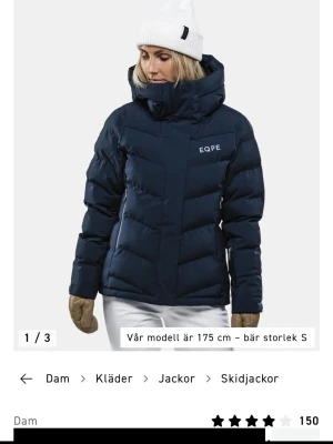 Mörkblå dunjacka från EQPE - Snygg mörkblå dunjacka från EQPE med hög krage och justerbar huva. Jackan har ett quiltat mönster, två sidofickor med dragkedja och logotyp på bröstet. Perfekt för kalla vinterdagar och skidåkning och håller dig varm med sitt tjocka dunmaterial.