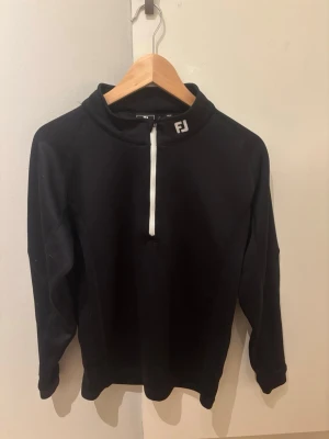 Svart halvzip tröja från FootJoy - Snygg svart långärmad tröja från FootJoy med halv dragkedja framtill och hög krage. Perfekt för lager på lager och sportig stil. Diskret logga på kragen och mjukt material som känns bekvämt hela dagen. Storleken är XL i barn och passar ca 160-170 cm i längd!