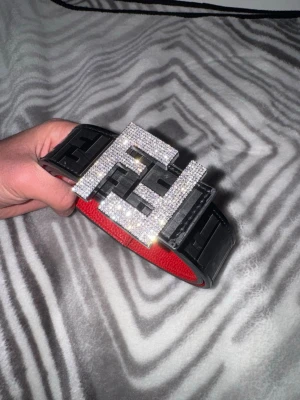 Svart skärp med glittrigt Fendi-spänne - Säljer ett svart skärp från Fendi med ett stort, glittrigt spänne format som Fendi-loggan. Skärpet har en bred design och insidan är röd. Perfekt för att lyfta din outfit med en exklusiv touch.