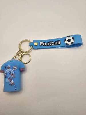 Blå nyckelring med fotbollstema - Nyckelring i silikon med blått band och texten 'Football'. Hängande tröja med tryck av en fotbollsspelare, nummer 9 och namnet Haaland. Guldiga metalldelar och fotbollsmotiv på bandet. Perfekt accessoar för fotbollsfans.