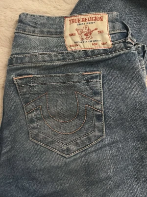 Blå jeans från True Religion - Snygga blå jeans från True Religion med rosa söm. Jeansen har låg midja. En del defekter som syns på bilderna. Storlek 25