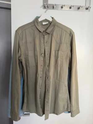 Beige skjorta från ASOS DESIGN - Stilren beige skjorta från ASOS DESIGN med klassisk krage, bröstfickor och knappar framtill. Skjortan är långärmad och har en avslappnad passform, perfekt för en trendig och enkel look.