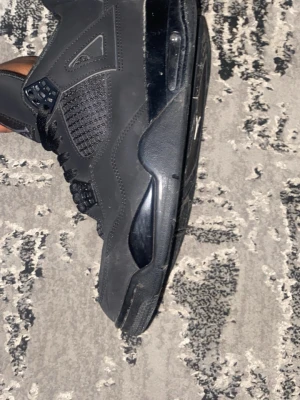 Svarta Air Jordan 4 Retro sneakers - Säljer ett par helsvarta Air Jordan 4 Retro sneakers med meshpaneler, synlig Air-enhet i sulan och klassiska Jumpman-loggan på tight plös. Skorna, i high-top modell, har svarta skosnören och detaljer i plast och mocka för en clean och sportig look.