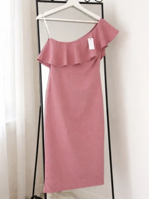 Rosa oneshoulder klänning med volang -  Helt ny rosa klänning med off shoulder-design och volang upptill. Har en smal passform och dragkedja i ryggen för enkel påklädning. Aldrig använd och har kvar originaltagg. Perfekt till sommar, fest eller middag. Är i storlek 40 men kan användas av mindre storlekar då den är väldigt tajt