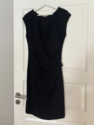 Svart klänning Tiger of Sweden  - Stilren svart klänning med draperad detalj framtill och kort ärm. Klänningen har en smickrande passform och är knälång. 