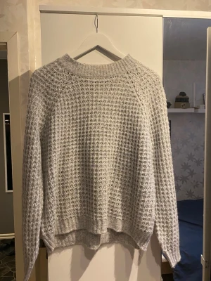 Ljusgrå stickad tröja 157 - Ljusgrå stickad tröja från 157 med grovstickad struktur och rund halsringning. Tröjan har långa ärmar och ribbade muddar vid halsen. Perfekt för lager på lager och en avslappnad look.