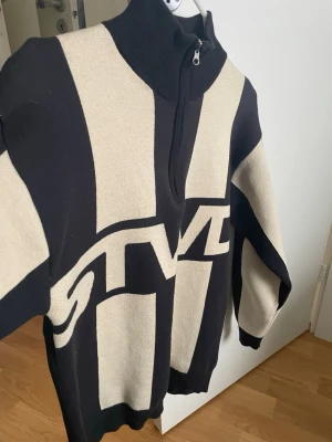 Svart-beige stickad tröja med zip - Stickad tröja i svart och beige med breda blockränder och STVL-tryck framtill. Hög krage med dragkedja och ribbstickade detaljer. Perfekt för dig som gillar streetwear och vill ha en unik look.