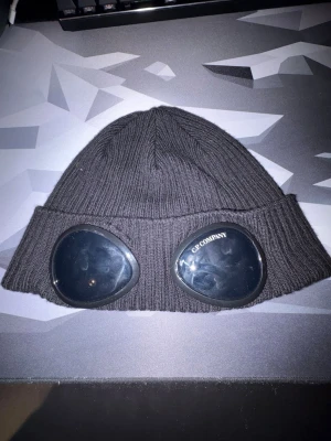 Svart mössa med goggles från C.P. Company - Säljer en svart ribbstickad mössa från C.P. Company med ikoniska inbyggda goggles framtill. Mössan är tillverkad i mjukt material och har en uppvikt kant. Perfekt för dig som vill ha en streetwear-look med edge.
