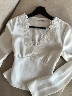Vit blus med spetsdetaljer från NA-KD - Supersnygg vit blus från NA-KD med v-ringning och broderade spetsdetaljer runt halsen.💗💗 Bl. Materialet är glansigt, perfekt för att lyfta vilken outfit som helst. Storlek 32.  Fri frakt fram tills fredag