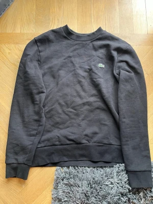 Svart tröja från Lacoste - Klassisk svart sweatshirt från Lacoste med rund halsringning och det ikoniska gröna krokodilmärket broderat på bröstet. Tröjan har ribbade muddar vid ärmslut och nederkant. Perfekt för en clean och stilren look.