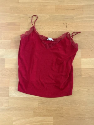 Röd ribbad linnetopp med spets i L från H&M - Snygg röd linnetopp med tunna axelband och v-ringning dekorerad med spets. Toppen har en ribbad struktur och är perfekt för lager på lager eller som statement-plagg. Enkel och trendig design som passar till många olika stilar.