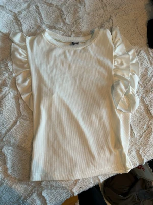 Vit ribbad topp med volangärm Shein - Säljer en vit ribbad topp från Shein med volangdetaljer längs axlarna. Toppen har rund halsringning och långa ärmar. Perfekt för dig som gillar en enkel men ändå söt stil.