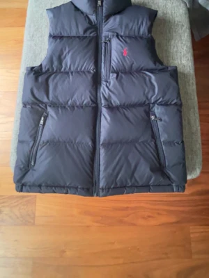 Mörkblå dunväst från Polo Ralph Lauren - Snygg mörkblå dunväst från Polo Ralph Lauren med dragkedja framtill och två praktiska fickor med dragkedja. Västen har en diskret röd logga på bröstet och är perfekt för lager-på-lager-stil. Tillverkad i ett glansigt material som ger en modern look.