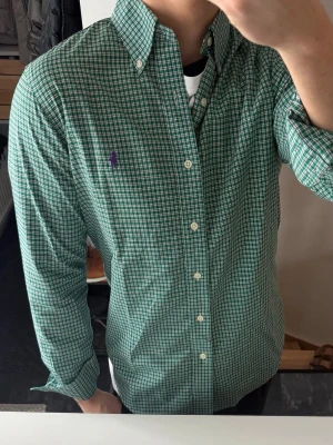 Grön rutig skjorta från Ralph Lauren - Säljer en grön och vit rutig skjorta från Ralph Lauren med klassisk button-down krage och lila broderad logga på bröstet. Skjortan har långa ärmar, vita knappar och är tillverkad i mjuk bomull. Perfekt för en clean och stilren look.