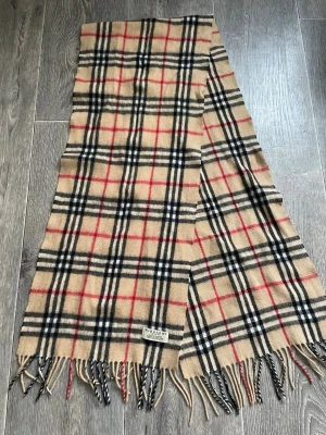 Beige rutig halsduk från Burberry - Klassisk beige halsduk från Burberry i mjuk ull med det ikoniska rutmönstret i svart, vitt och rött. Halsduken har tvinnade fransar i ändarna och en vävd Burberry-etikett. Perfekt accessoar för att lyfta din outfit.