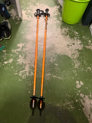 Orangea skidstavar från Leki - Säljer ett par orangea skidstavar från Leki med svarta handtag och handledsremmar. Stavarna har rund truga och spets i metall, perfekta för vintersport och skidåkning. Snygg och sportig design som sticker ut i backen. Nypris 800