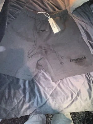 Svarta Essentials shorts med snörning - Säljer ett par svarta shorts från Fear of God Essentials med broderad logga på benet och justerbar snörning i midjan. Sköna och stilrena, perfekta för chill dagar eller streetwear-stil. Materialet känns mjukt och Iyxigt.