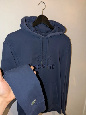 Blå hoodie från Lacoste med broderad logga - Mörkblå hoodie från Lacoste med broderad krokodil och text på bröstet. Klassisk huva med dragsko och känguruficka framtill. Ärmslutet har en liten broderad Lacoste-logga.  100% bomull, storlek m orginal pris 1250kr