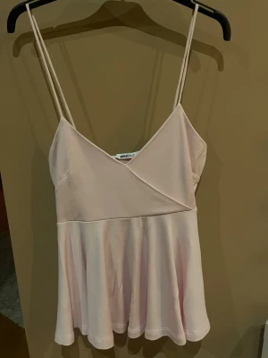 Ljusrosa linne från Gina Tricot - Supersött ljusrosa linne med tunna axelband och v-ringning från Gina Tricot. Toppen har en omlott-detalj framtill och ett flowy, utsvängt fall nedtill. Perfekt för en trendig och feminin look.