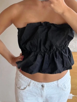 Svart tubtopp med volang - Svart axelbandslös tubtopp med volangkant nedtill och rynkad midja. Perfekt för en trendig och avslappnad look. Toppens material känns lätt och luftig.