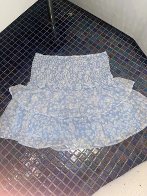 Blå volangkjol från Lindex - Supersöt blå kortkjol från Lindex med vita blommor och volangdetaljer. Kjolen är perfekt att ha till sommaren. TAR GÄRNA EMOT PRISFÖRSLAG💗