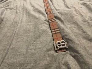 Beige rutigt bälte med BB-spänne - Snyggt bälte från Burberry i klassiskt beige och röd-rutigt mönster. Bältet har ett stilrent BB-spänne i silverfärg och är tillverkat i skinn. Perfekt accessoar för att lyfta din outfit.